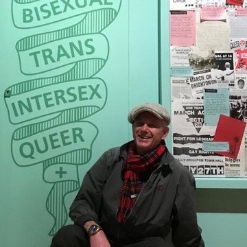 Man in flat cap and red tartan scarf crouched in front of turquoise museum wall with signage from queer exhibition.