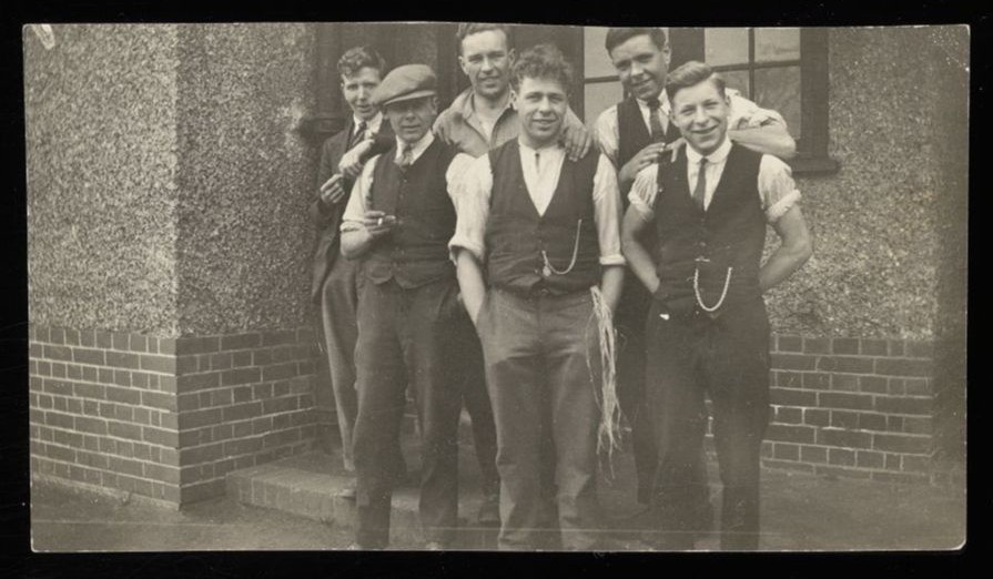 Informal photograph of a small group of men in waistcoats and shirtsleeves outside a building.