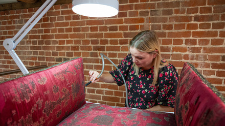 Woman cleaning an old red silk sofa with a tiny brush on a long hose.