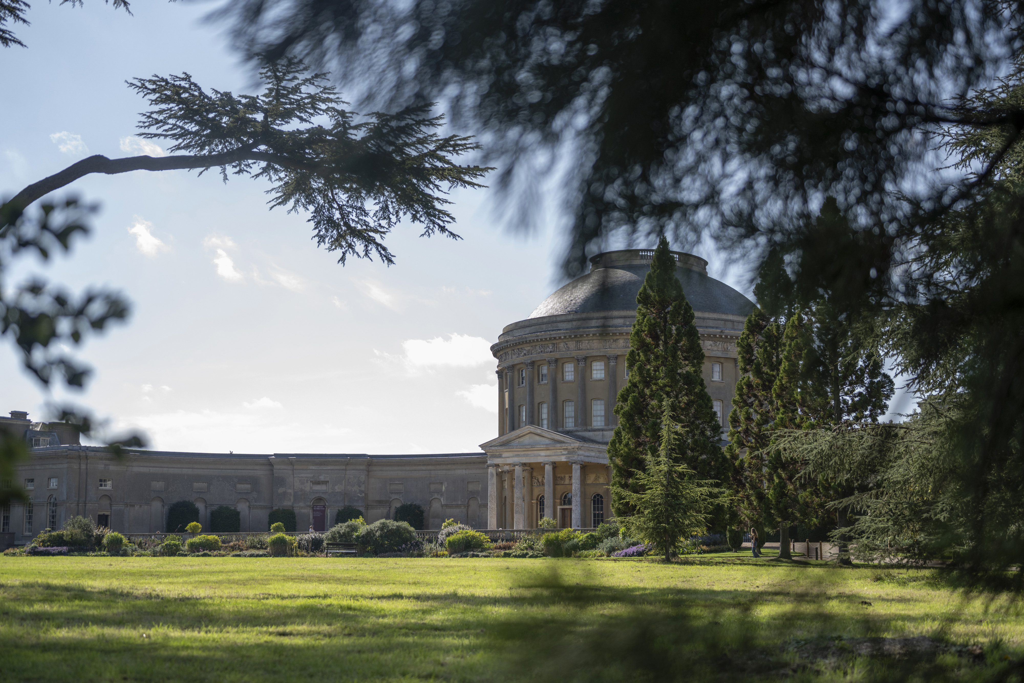 Grand round, stone house with long wing extending away from it, set in green parkland.