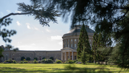 Grand round, stone house with long wing extending away from it, set in green parkland.