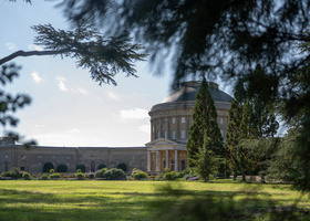 Grand round, stone house with long wing extending away from it, set in green parkland.