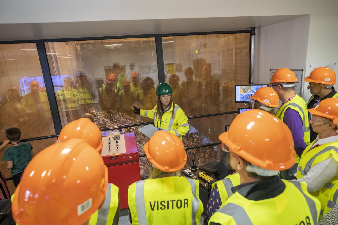 A group of visitors with their back to us, wearing hi-vis jackets being shown how the waste is gattered through a glass window.