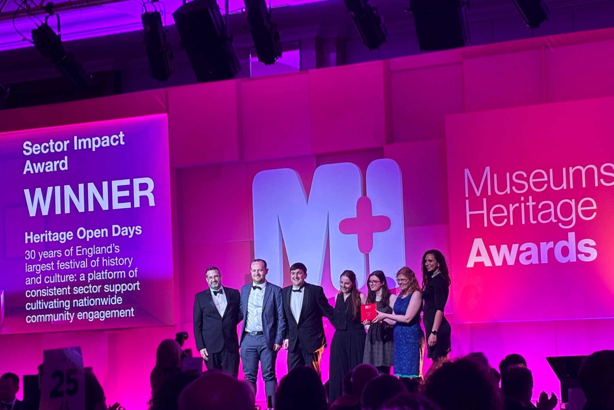 7 people stood on a pink lit stage receiving an award.