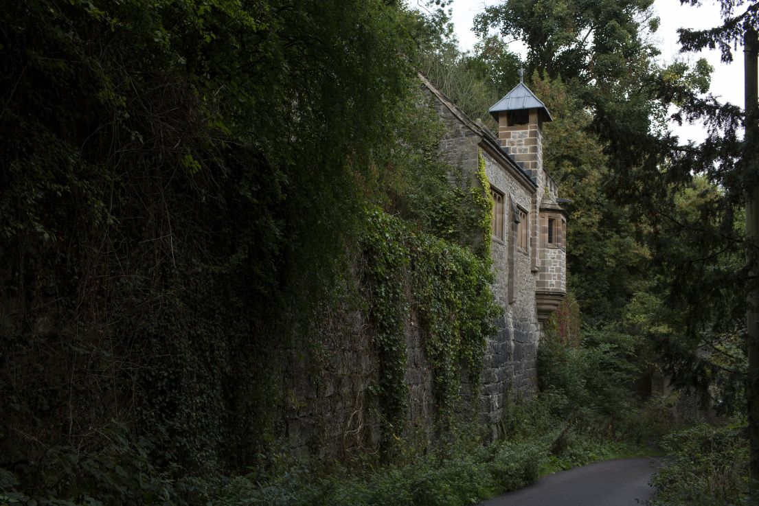 A church built into the rocks of a hillside. The high walls of the church run against a narrow road, the walls overgrown with plants and trees.