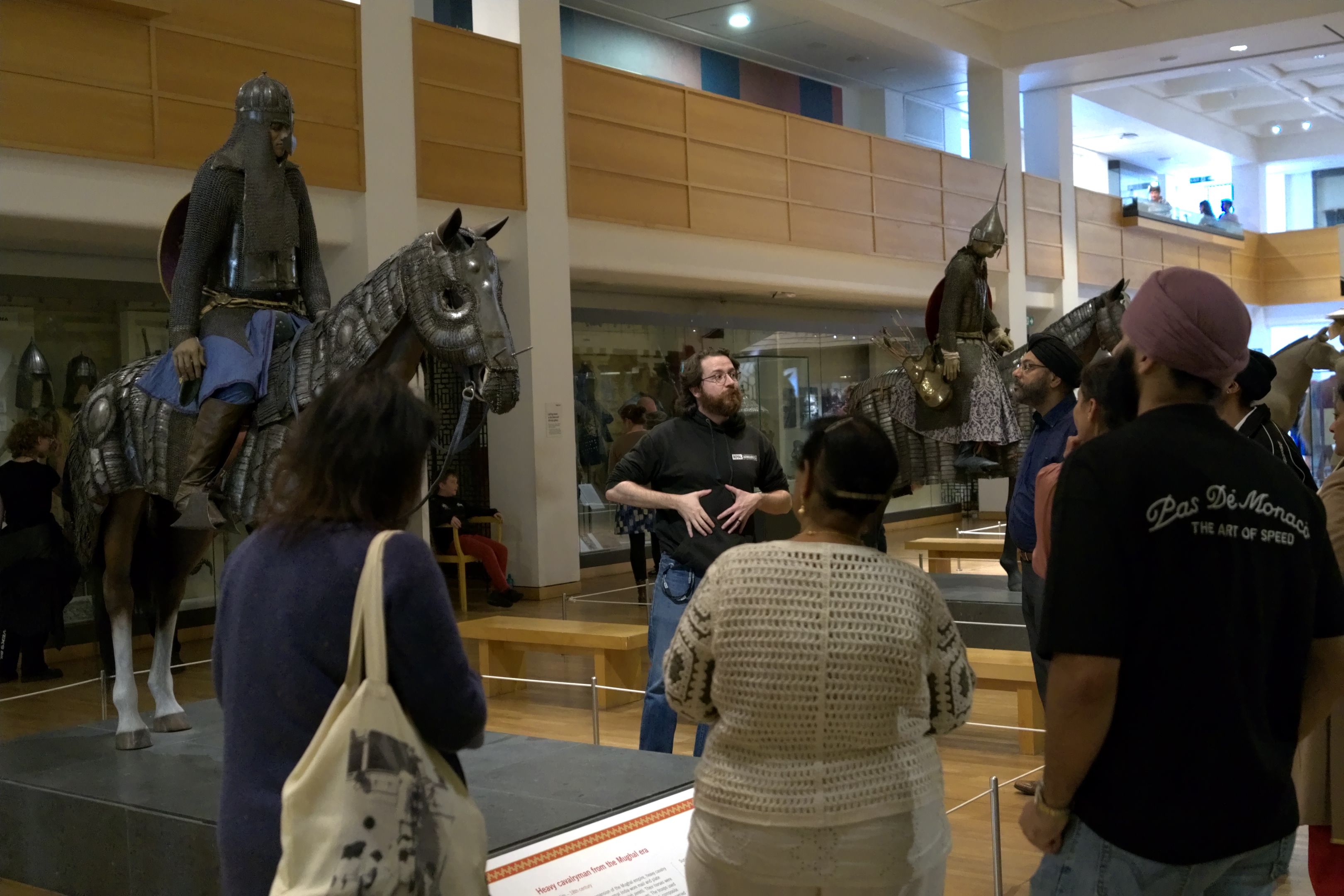 Bearded white man talking to a group of Asian people in a gallery space next to a model of an armoured horse.
