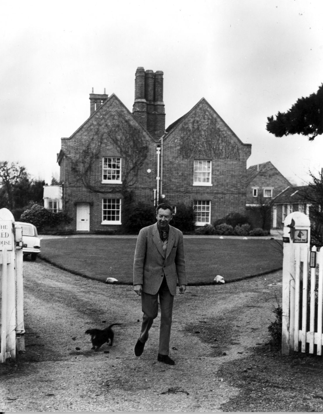 A black and white images of a man in a suit, looking towards the 17th-century farmhouse. Stood by a white fence, behind him the driveway.