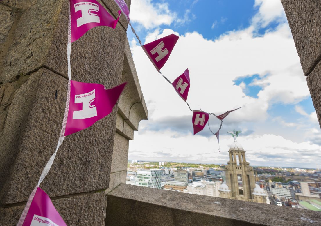 The Pink HODs bunting attached on an open balcony on a stone building, looking across to the Liverpool skyline and the metal statue of a liver bird.