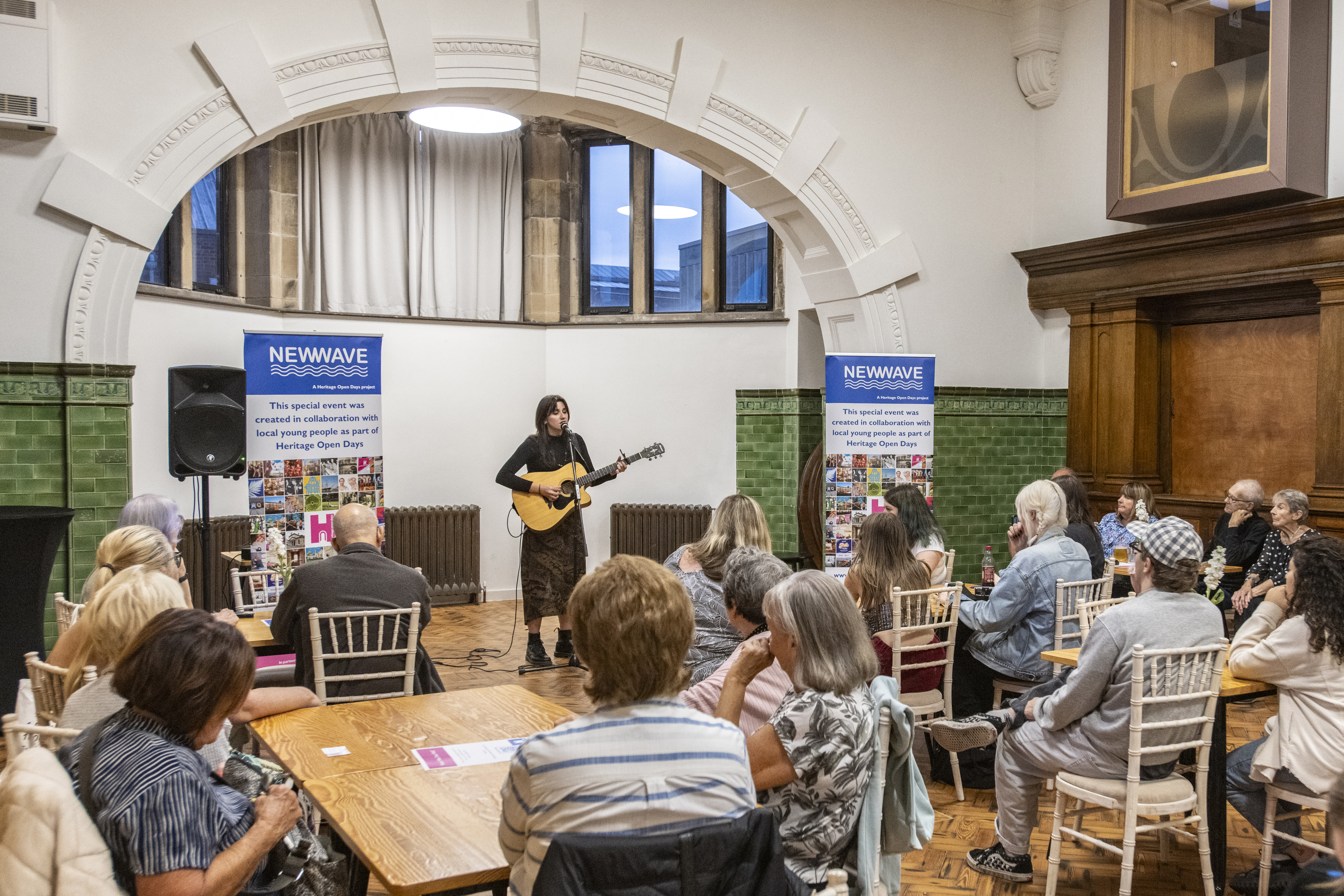 A small crowd sat around tables watching a performer with a guitar.