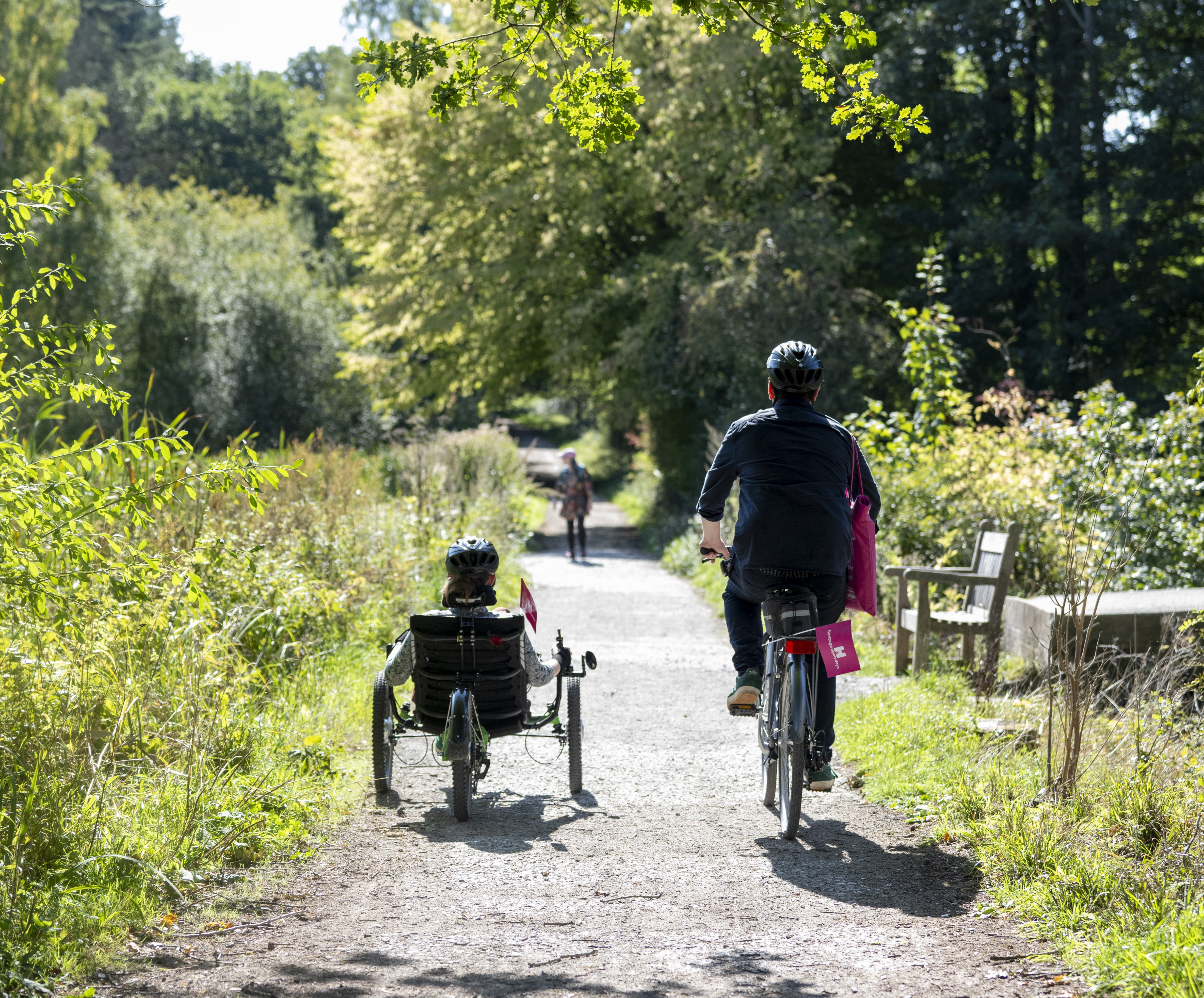 Two people cycling down a trail bordered by trees and plants. One on a recumbent tricycle.