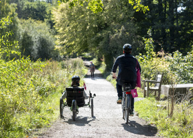 Two people cycling down a trail bordered by trees and plants. One on a recumbent tricycle.