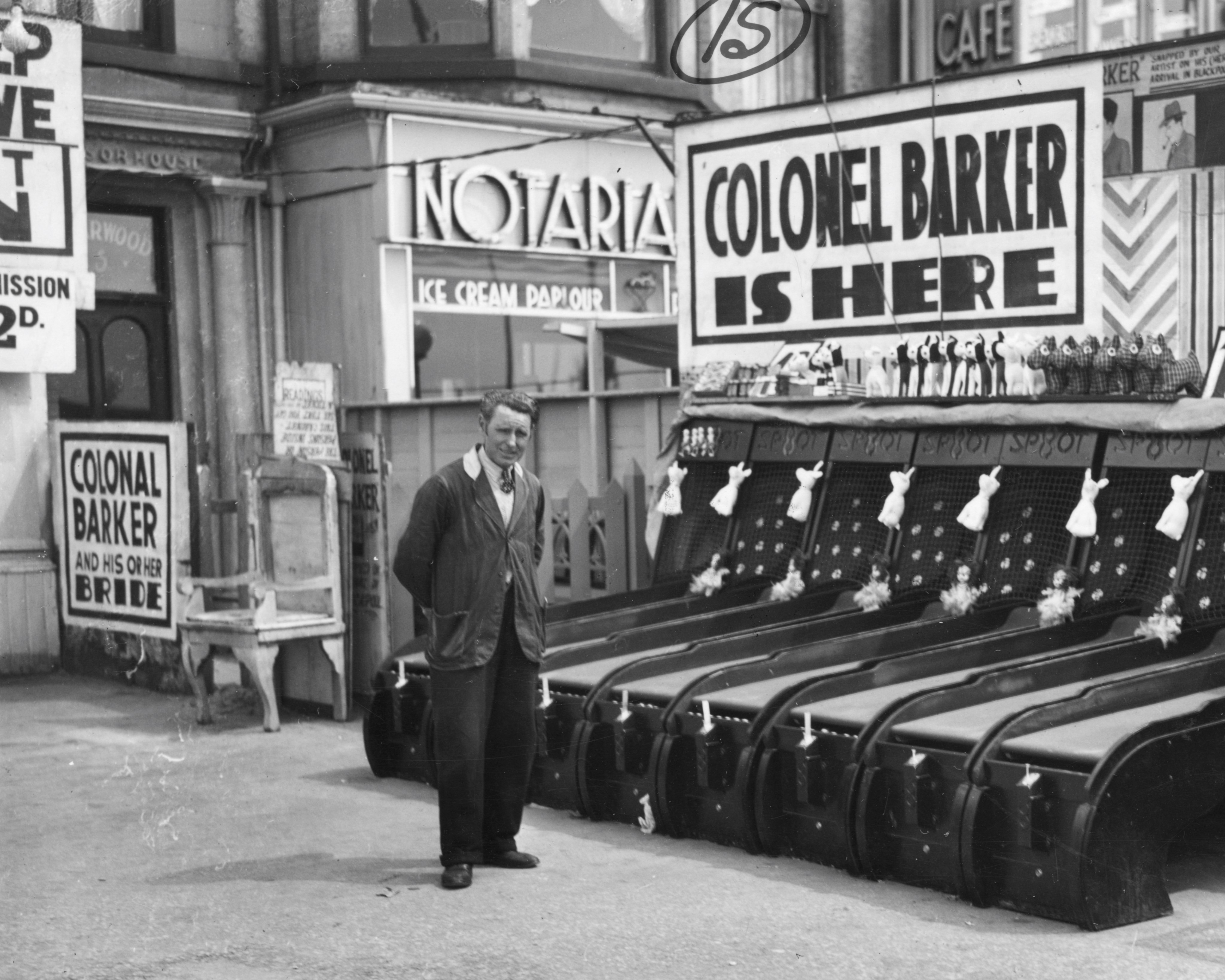 Man stood in front of arcade amusement game near an ice cream parlour with large adverts for Colonel Barker.