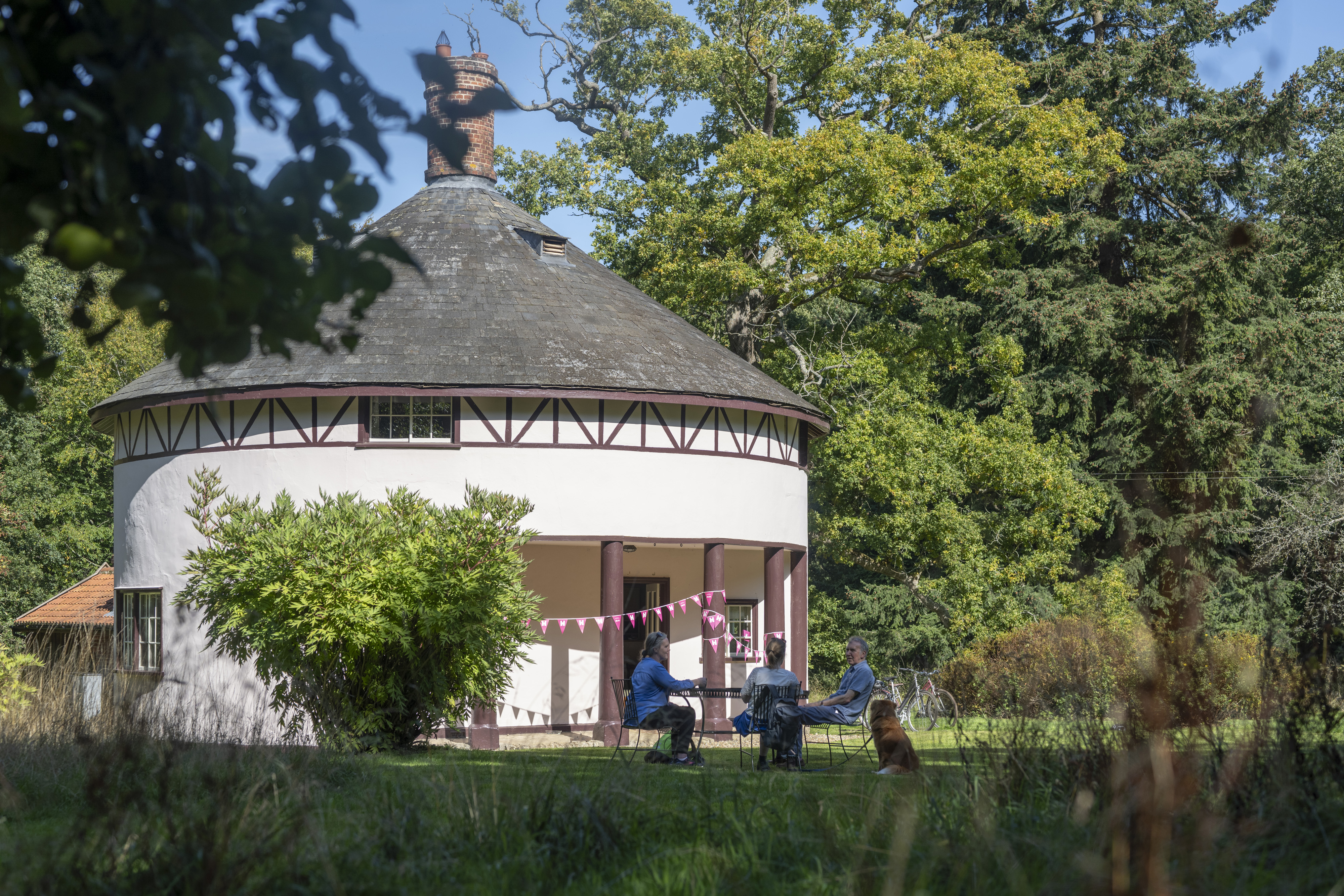 People sat in garden outside a small whitewashed round house with conical roof decorated with pink bunting.
