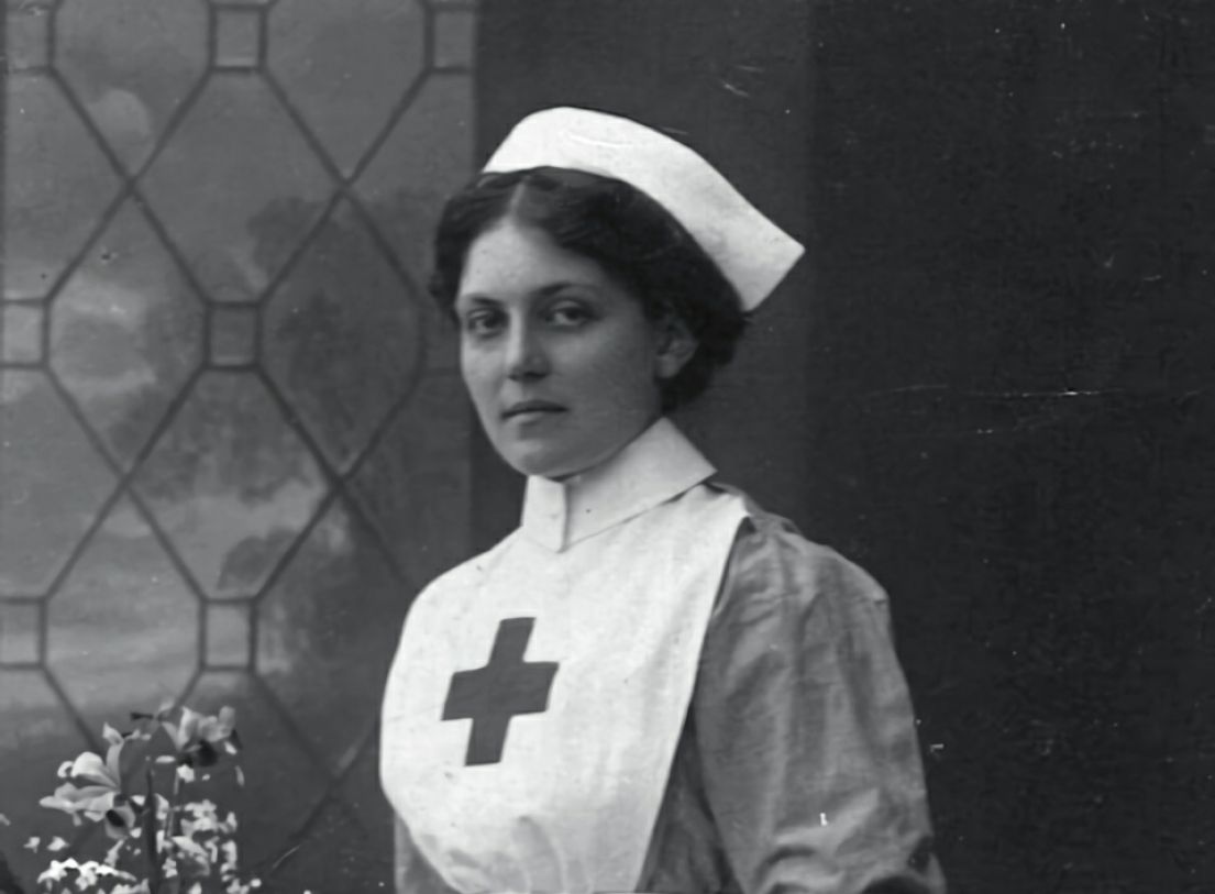 A cropped black and white image of a women stood by a window, she is dressed in an Edwardian nurse, which a crisp white hat and apron with a red cross