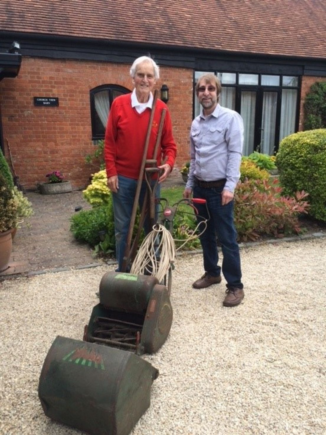 Two men, one wearing a red jumper and jeans, the other in a long sleeved checked shirt and jeans standing behind an old and slightly rusted lawnmower.