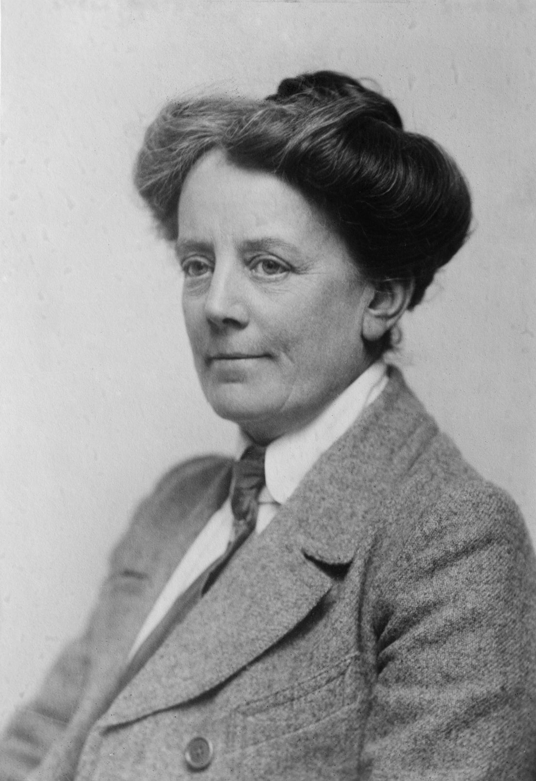 A black and white headshot of a women. Her long hair tied up into a bun, she wears a tidy suit jacket.