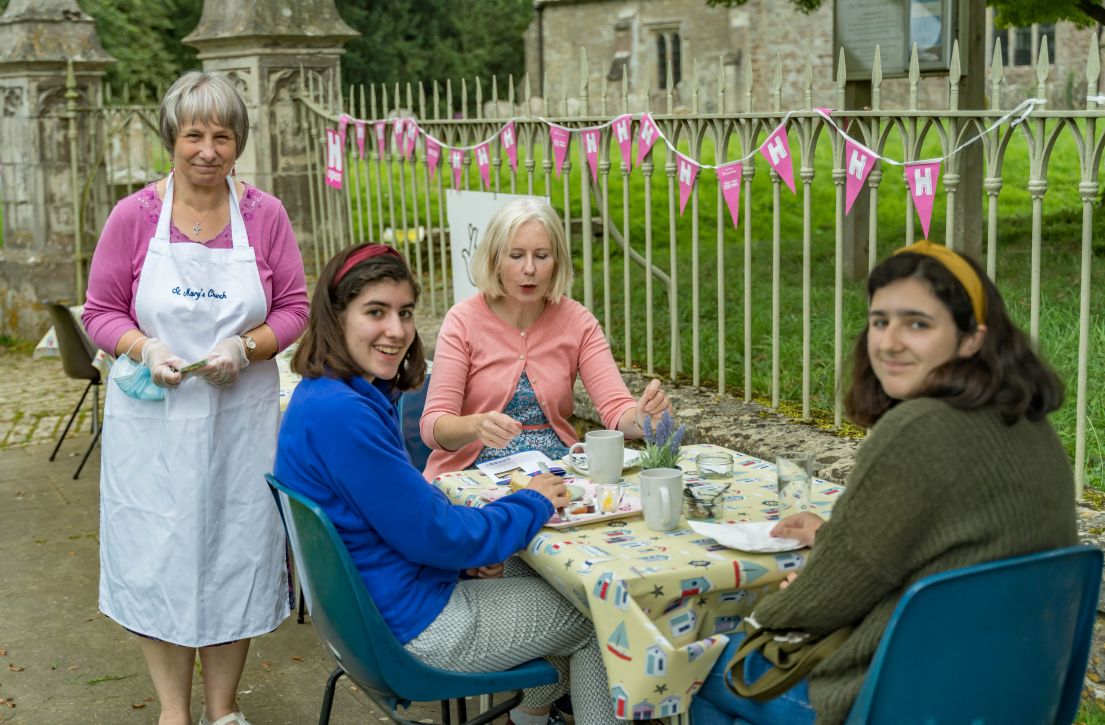 Four ladies sat on chairs around a clothed table enjoying afternoon tea. Behind them is HODs pink bunting,.