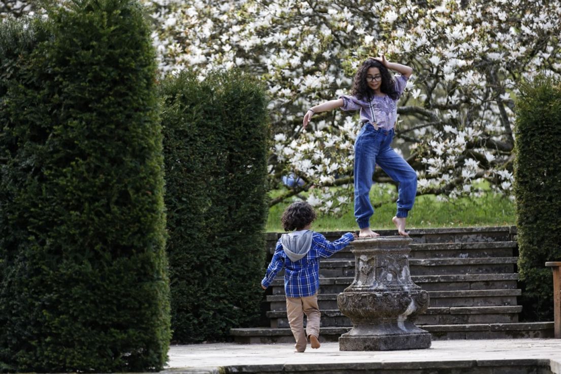 A girl with curly brown hair, purple shirt, suspenders and jeans striking a pose on a stone font in a garden. At the base is a small child.