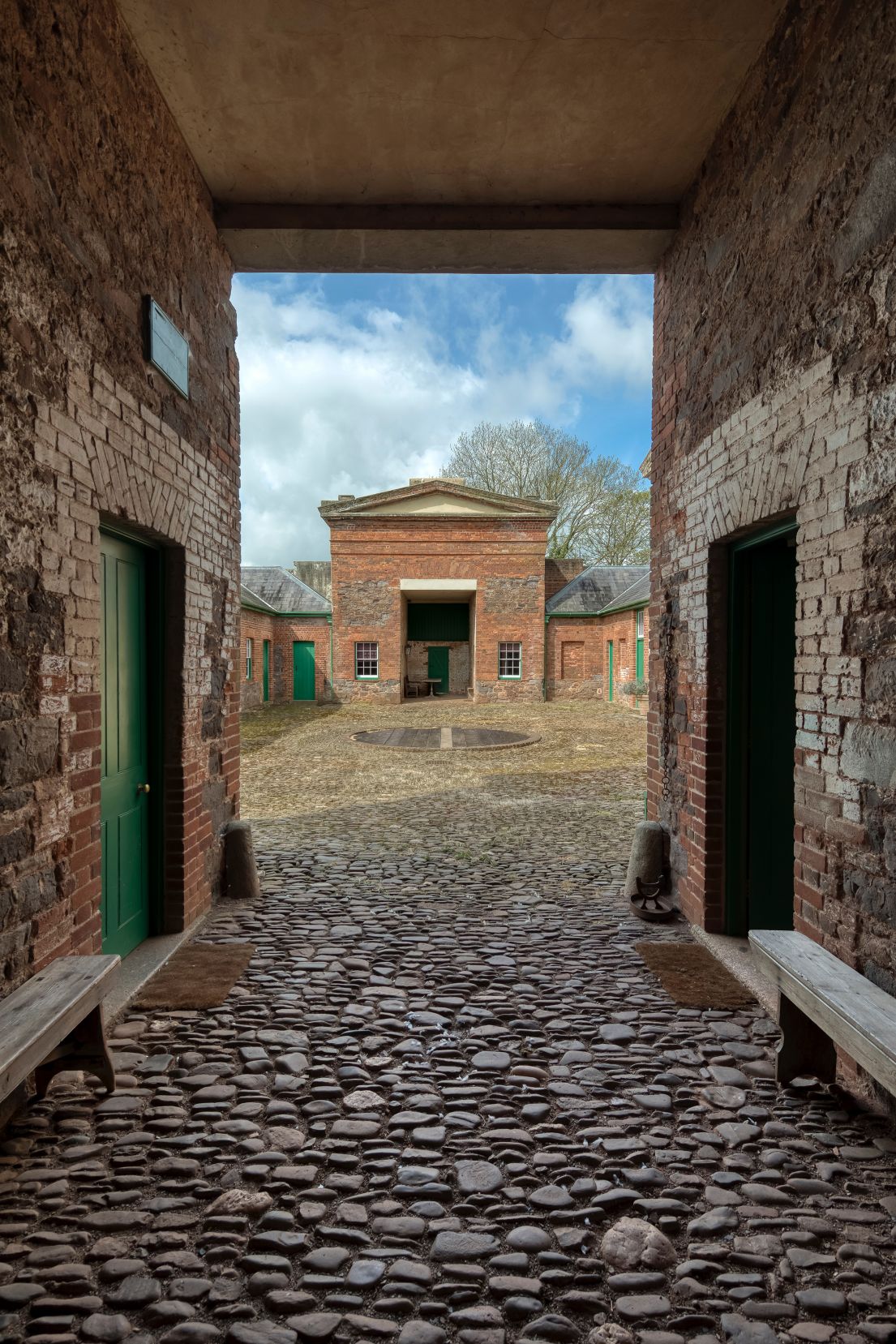 Looking through a cobbled passage towards a cobbled courtyard. Surrounding the courtyard are red bricked building with green doors.