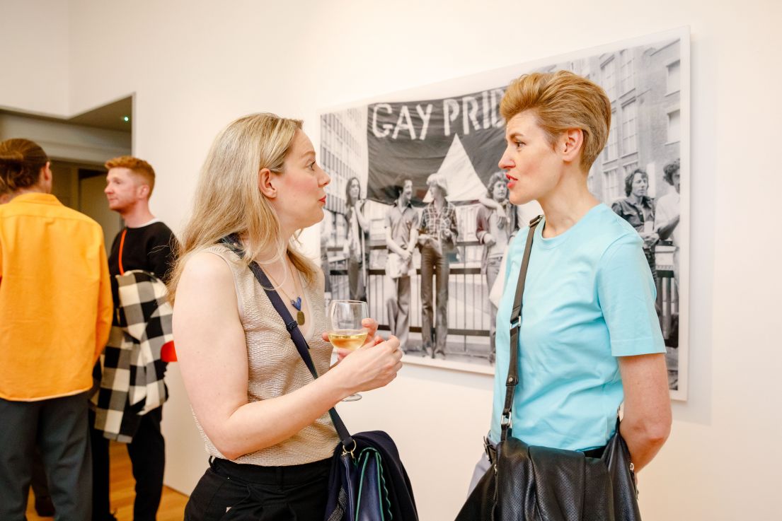 Two women having a discussion - behind them is a photograph of people standing by railings with a Gay Pride painted banner behind.