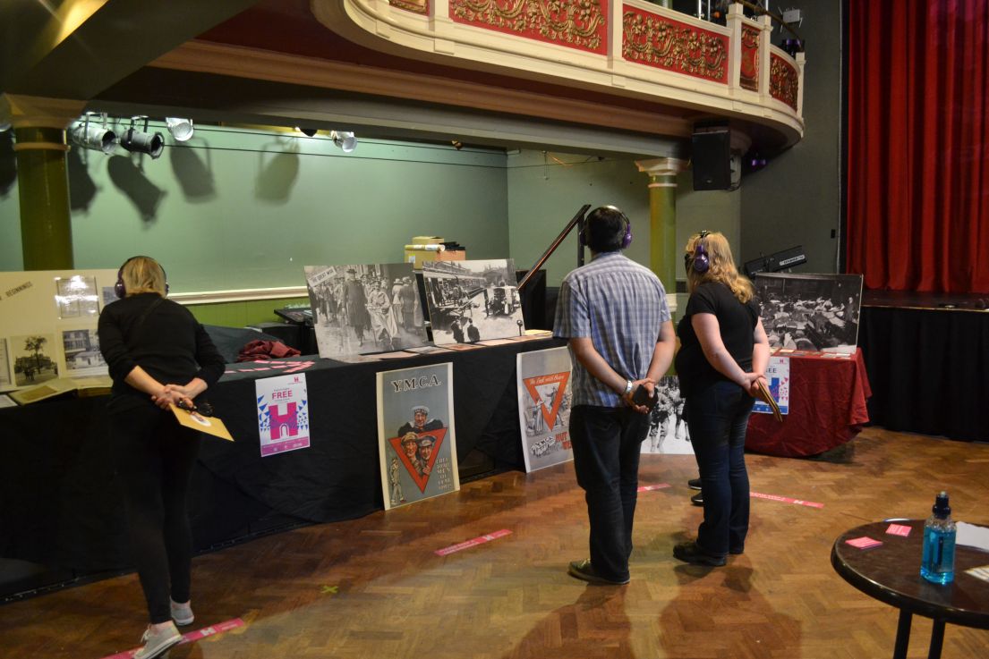 Three people, in a socially distanced fashioned, listening with headphones and looking at artefacts displayed on the theatre stage.