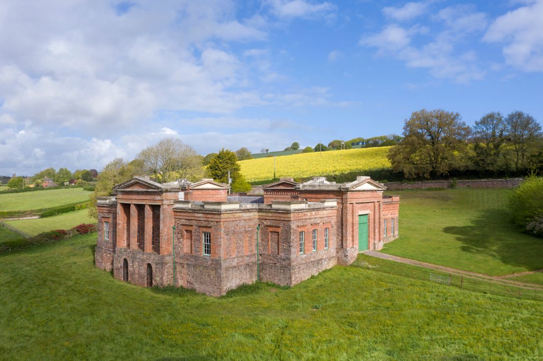 A drone photograph taken from a distance of a red bricked building in a field. The building has green accents of doors, windowpanes and gutters.