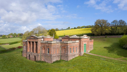 A drone photograph taken from a distance of a red bricked building in a field. The building has green accents of doors, windowpanes and gutters.