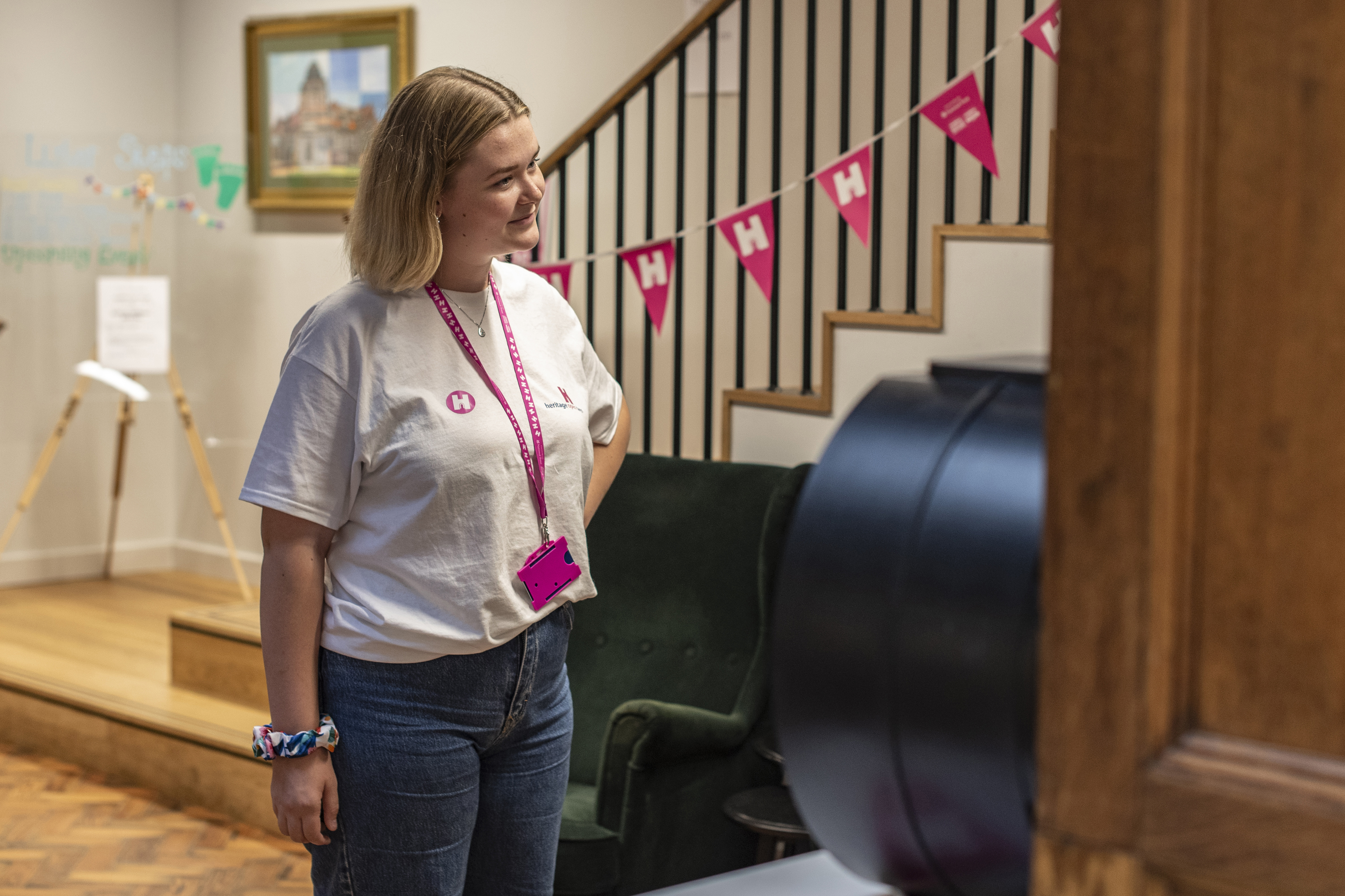 A lady wearing a white shirt with HODs logo, A HODs logo lanyard standing in a lobby with pink bunting running up the stairs