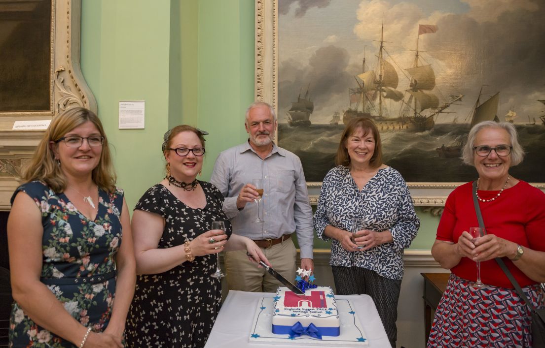 Five individuals with a glass of bubbles, standing around the HODs birthday cake. One lady is poised with a knife, ready to cut the cake.
