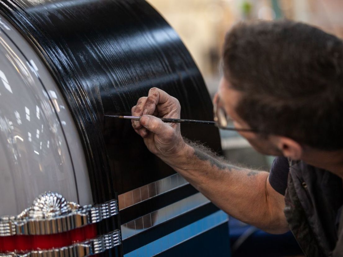 Man with short brown hair and glasses, carefully using a long paint brush to touch up the black paint on-top of a jukebox.