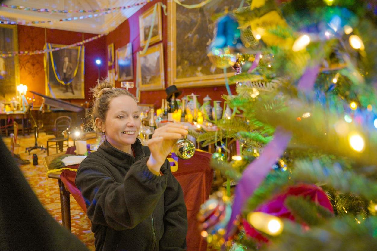 Woman hanging a bauble on a Christmas tree in a red walled room.