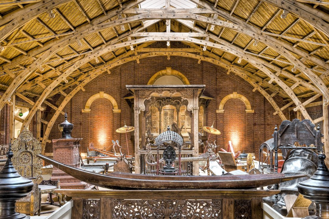 A large gallery with red brick walls and a wooden roof. A long, thin wooden boat is foregrounded and various Indian objects are on display.
