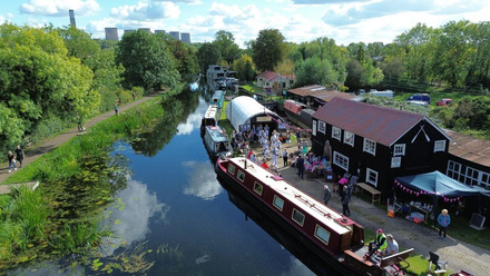 Aerial view of canal through green countryside with boat and buildings decorated in bunting in foreground.