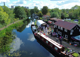 Aerial view of canal through green countryside with boat and buildings decorated in bunting in foreground.