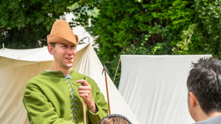 Man in cap and green coat holding an arrow in front of some white tents.