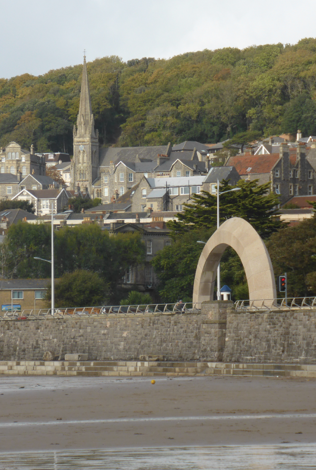 Looking from the beach, this looks up towards the stone wall, a arch of stone over an exit, with the town in the background with a large wood behind.