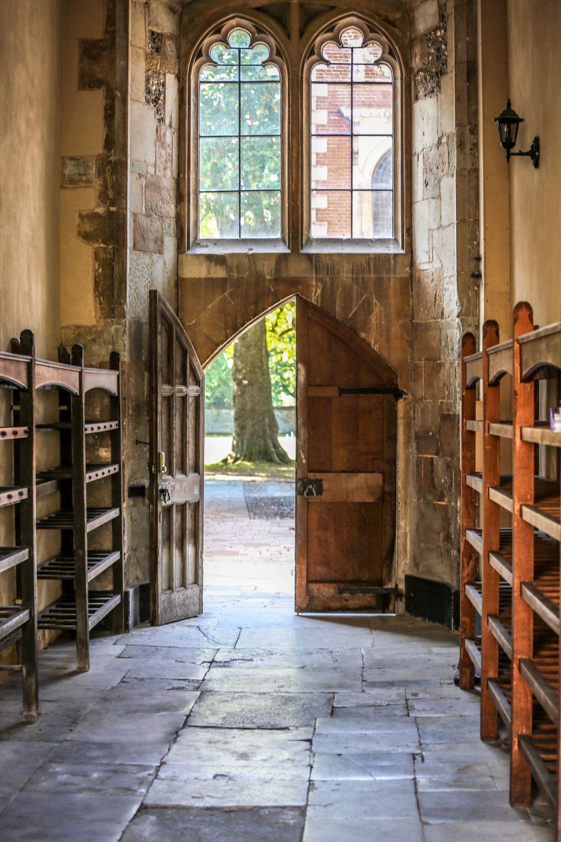 A stone floored hallway with deep brown crafted wooden shelves each side leading towards a stone archway with a double wooden door and windows above.