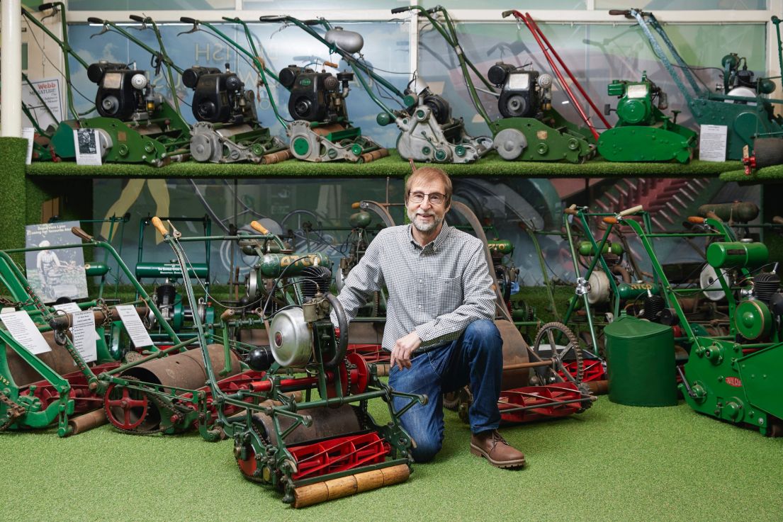 A man with short hair, wearing a checked shirt and jeans, kneeling down in-front a collection of various different metal green lawnmowers.