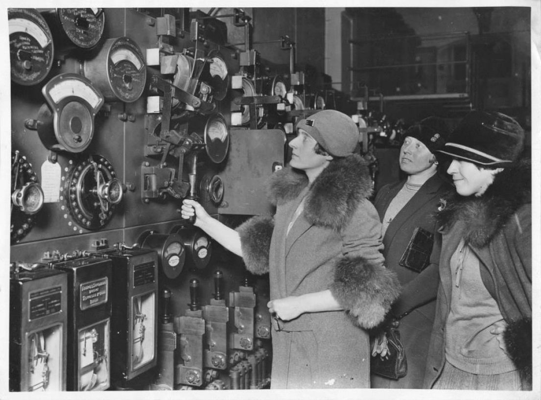 A black and white image of three women dressed in 1930's coats with fur trim and deep hats, examining dials in a factory.