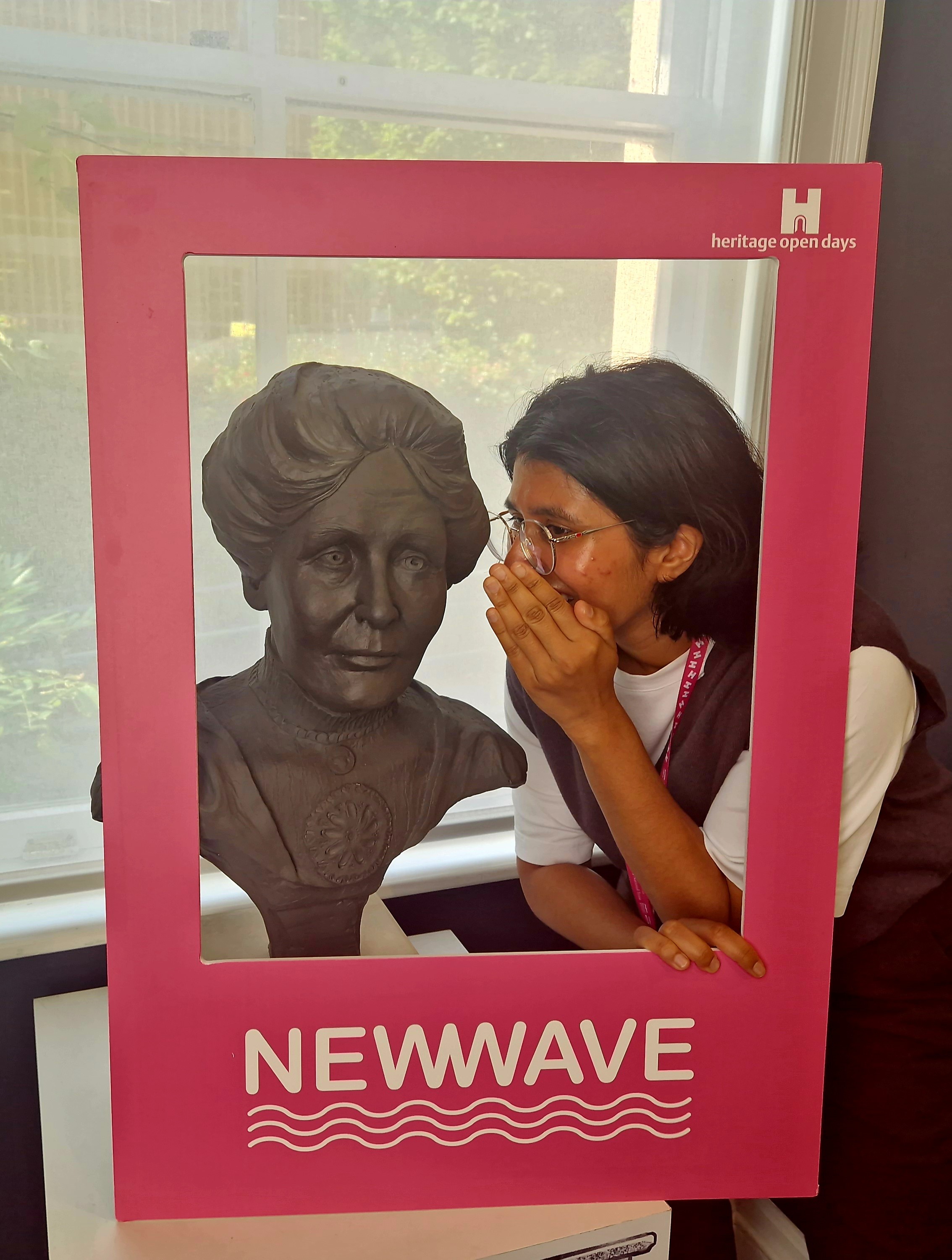 A person whispering to a bust of Emmeline Pankhurst within a pink New Wave selfie frame.
