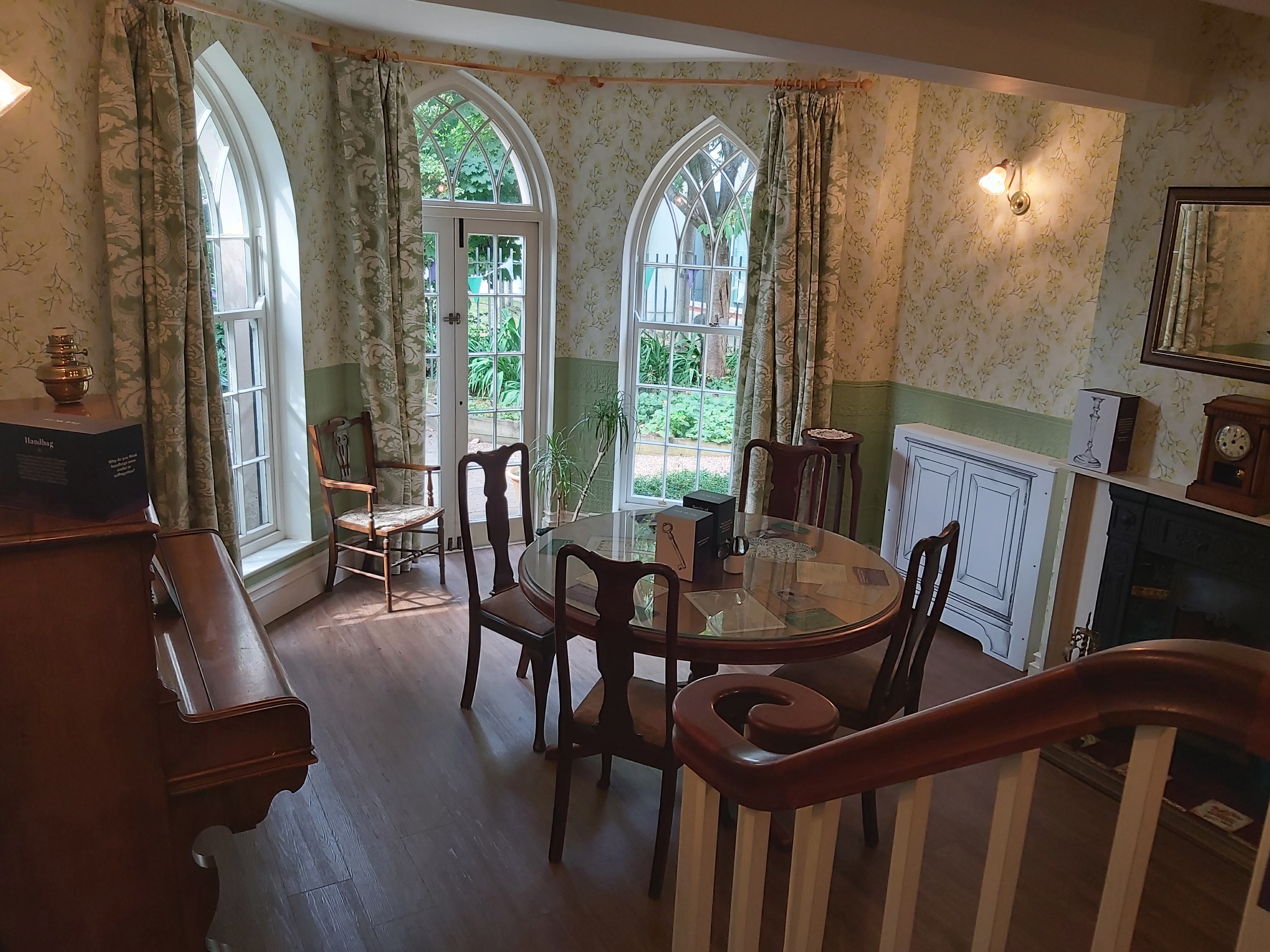 Floral wallpapered room with floor to ceiling windows lighting a small round table set with 4 chairs, and an upright piano against the wall.