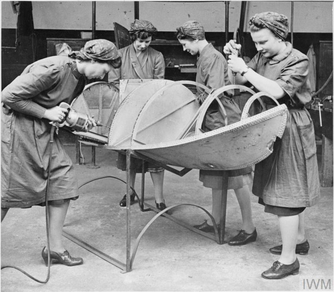 Four women in overalls and hair scarfs, bent over and working on structure of what would later become a seaplane float.