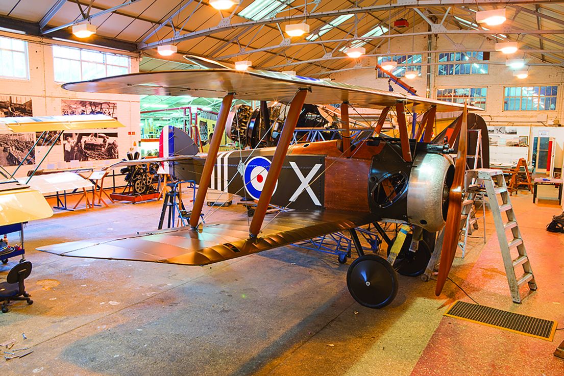 Inside an air hanger turned into a museum. A bi-wing wooden plane with a large propeller sites proud in the centre.