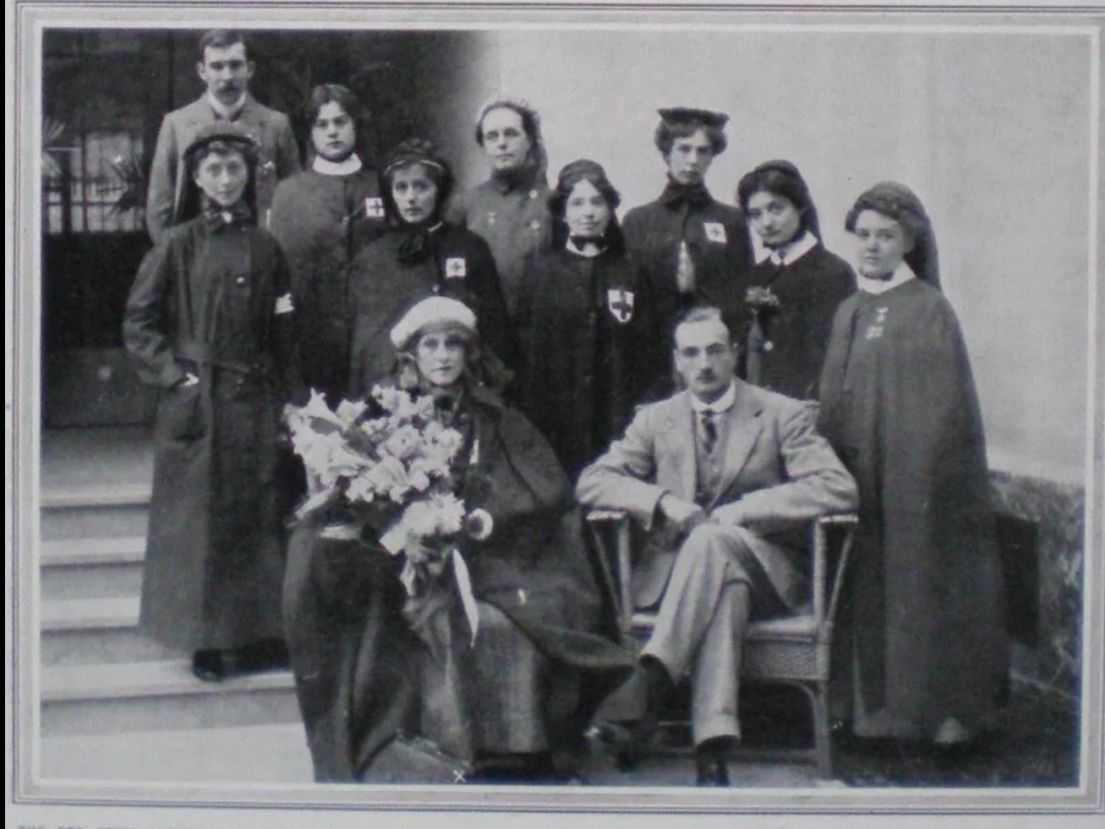 A black and white image of doctor and nurses in cape coats outside a building. Two of the individuals are sat and the others are gathered around on.