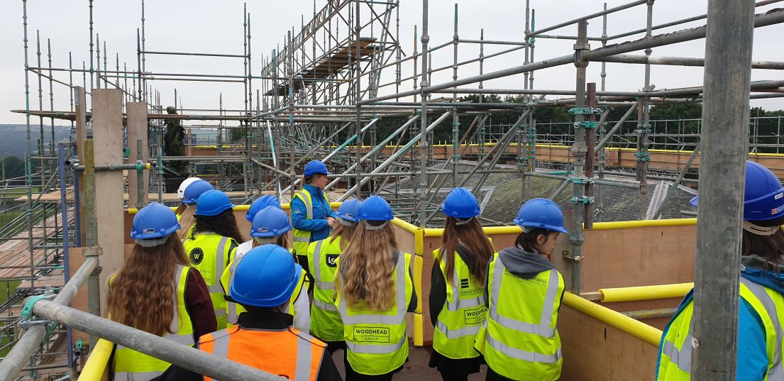 A group of girls being shown around the scaffolding of the roof conservation at a stately home. They all wear hi-vis jackets and hard hats.