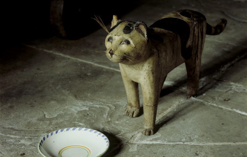 Wooden cat stands by china saucer on stone flagged floor.