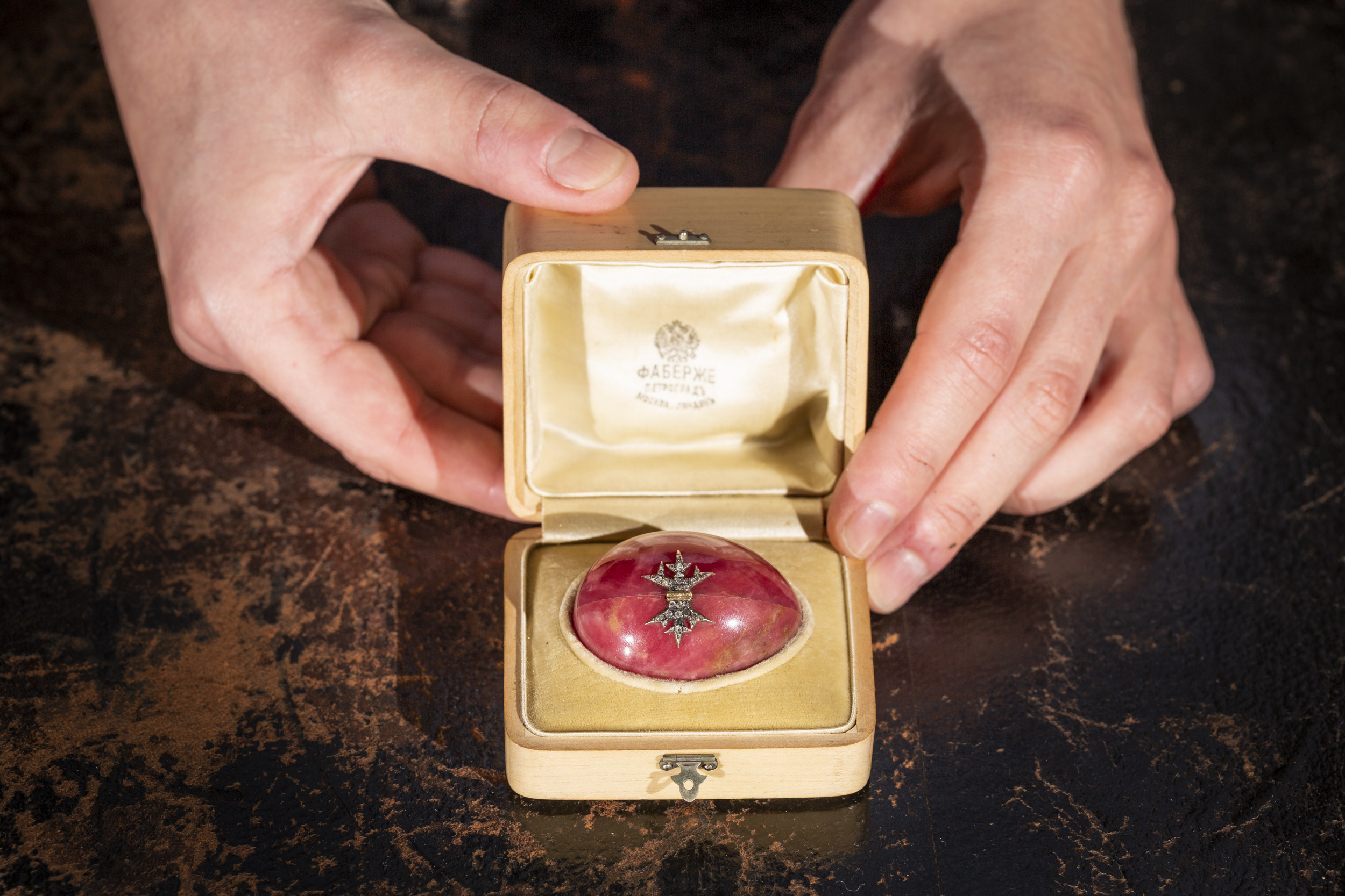 Two hands opening a small cream box containing a pink egg with a diamond snowflake on it.
