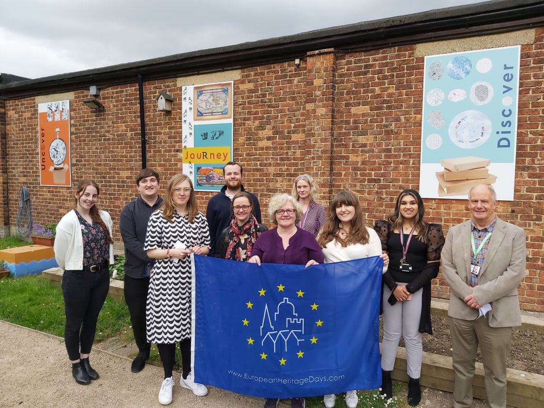 A group of people stood in-front of a small planting garden holding the European Heritage Days flag.