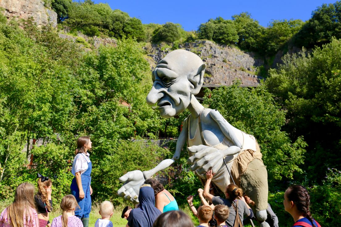 The 4 meter tall giant grey puppet which is controlled by three men, entertaining families and children in a field, with green trees in the background