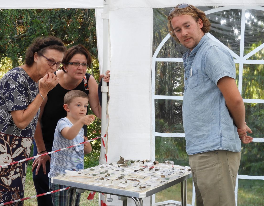 A women and child discussing artefacts that are laid on a table with a man in a blue short sleeved shirt. They are pointing at something not in view.